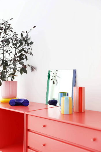 A modern coral sideboard displays the &klevering Ligne Vase, Set of 3, a pink plant pot with leafy branches, a wavy mirror, and stylish décor, all set against a crisp white wall.