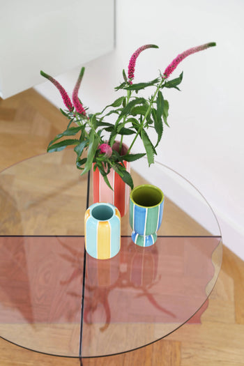 A glass table displays a pink vase with pink and green flowers beside pastel-striped &klevering Ligne Vases (Set of 3) by &klevering, creating a charming decor accent on a wooden floor.