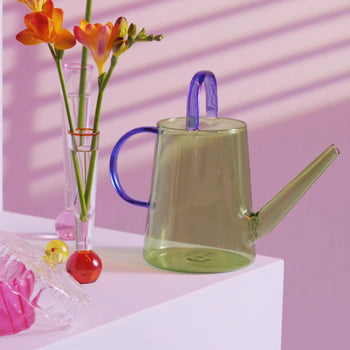 The &klevering Loop Watering Can by &klevering, featuring a translucent green glass body with a blue handle and lid, sits beside a vase of orange and yellow flowers—perfect for stylish plant care against a pink, striped-shadow background.