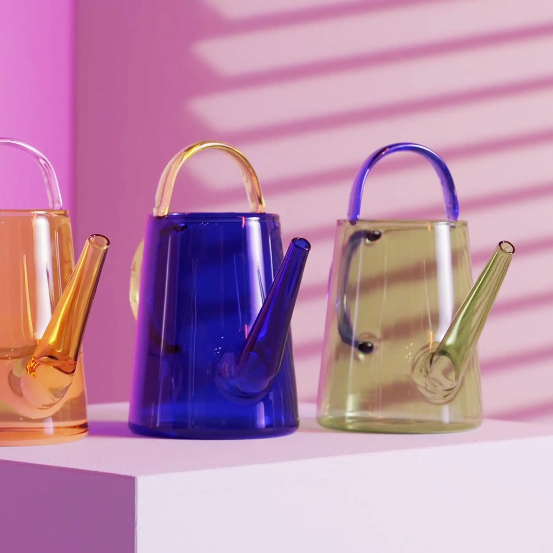 Three &klevering Loop Watering Cans with green glass bodies and blue handles are displayed on a white surface against a pink background—an inviting way to water your plants in style.