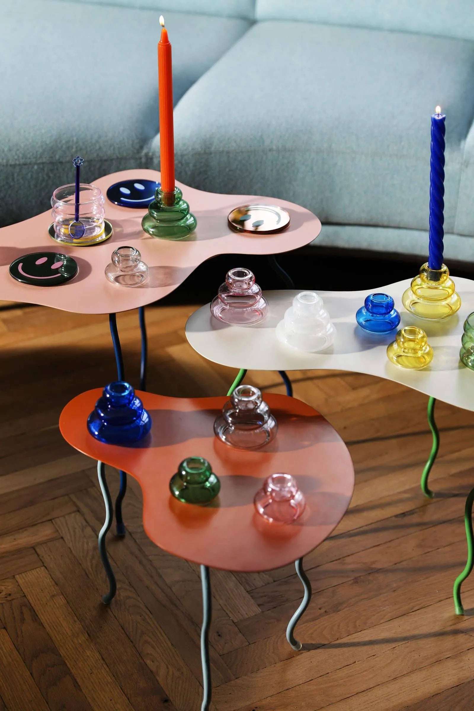 &klevering Marshmallow Candle, Set of 5 and colorful glass candle holders are displayed on three wavy tables with thin, curvy legs placed on a wooden floor next to a light blue sofa.