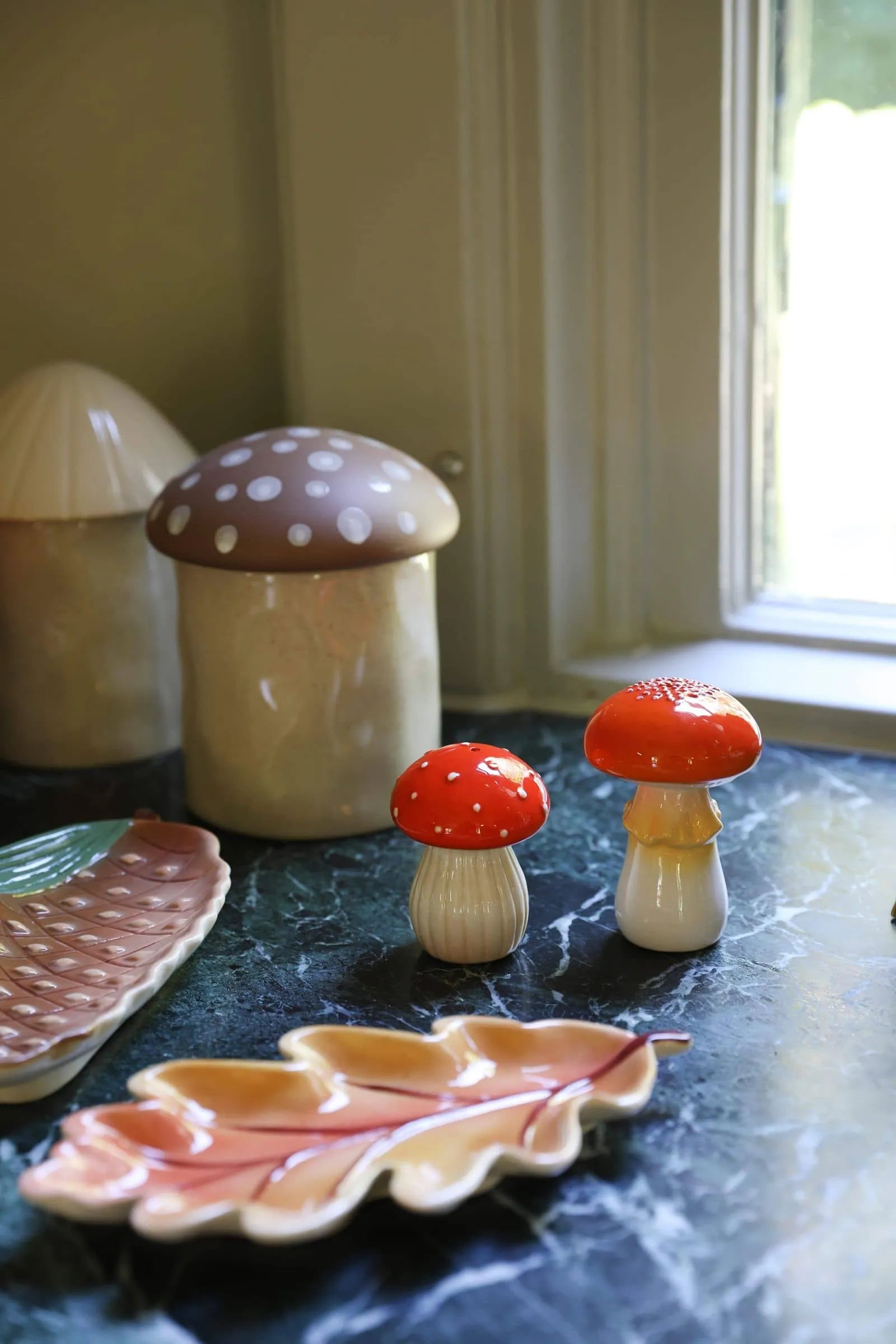 The &klevering Mushroom Salt & Pepper Set, featuring red and beige caps, sits on a green marble countertop by the window. Leaf-shaped ceramic dishes from &klevering add charm to this cozy kitchen’s dining experience.