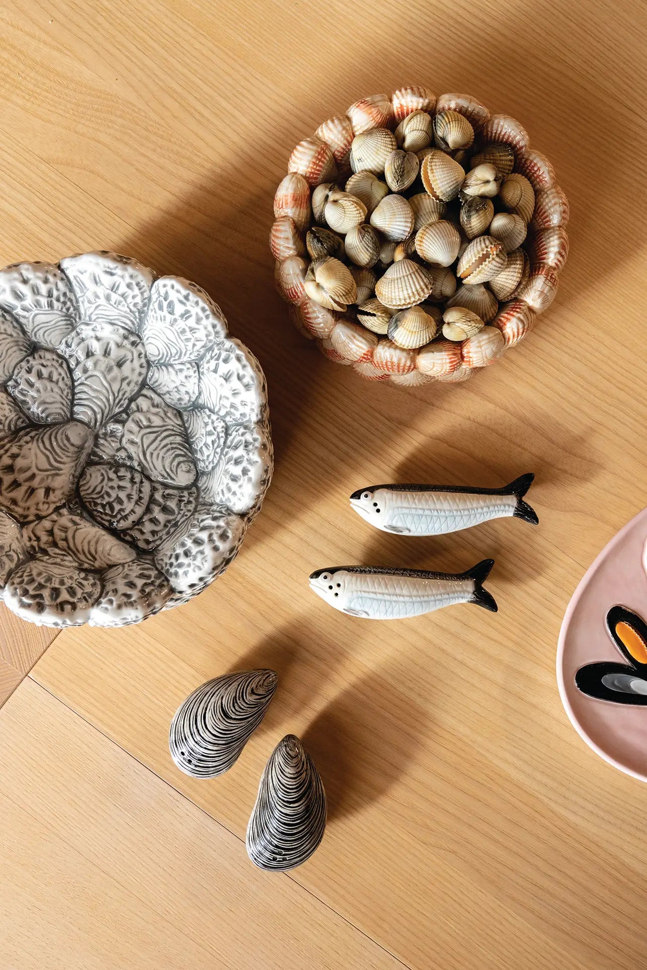 Ceramic shells, a bowl of seashells, two fish figurines, and the &klevering Oyster Bowl by &klevering with mussel designs are arranged on light wood—ideal for serving appetizers or delighting oyster lovers.