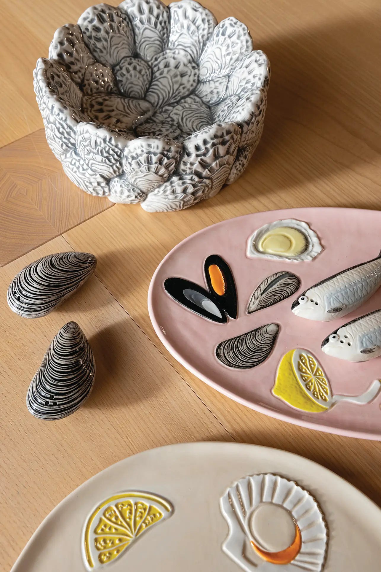 Ceramic dishware on a wooden table, including the detailed &klevering Oyster Bowl—perfect for appetizers—a pink plate with fish, mussels, and lemon designs, two mussel-shaped items, and a cream plate with lemon and shell motifs for oyster enthusiasts.