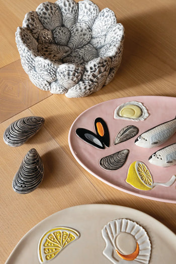 Ceramic dishware on a wooden table, including the detailed &klevering Oyster Bowl—perfect for appetizers—a pink plate with fish, mussels, and lemon designs, two mussel-shaped items, and a cream plate with lemon and shell motifs for oyster enthusiasts.