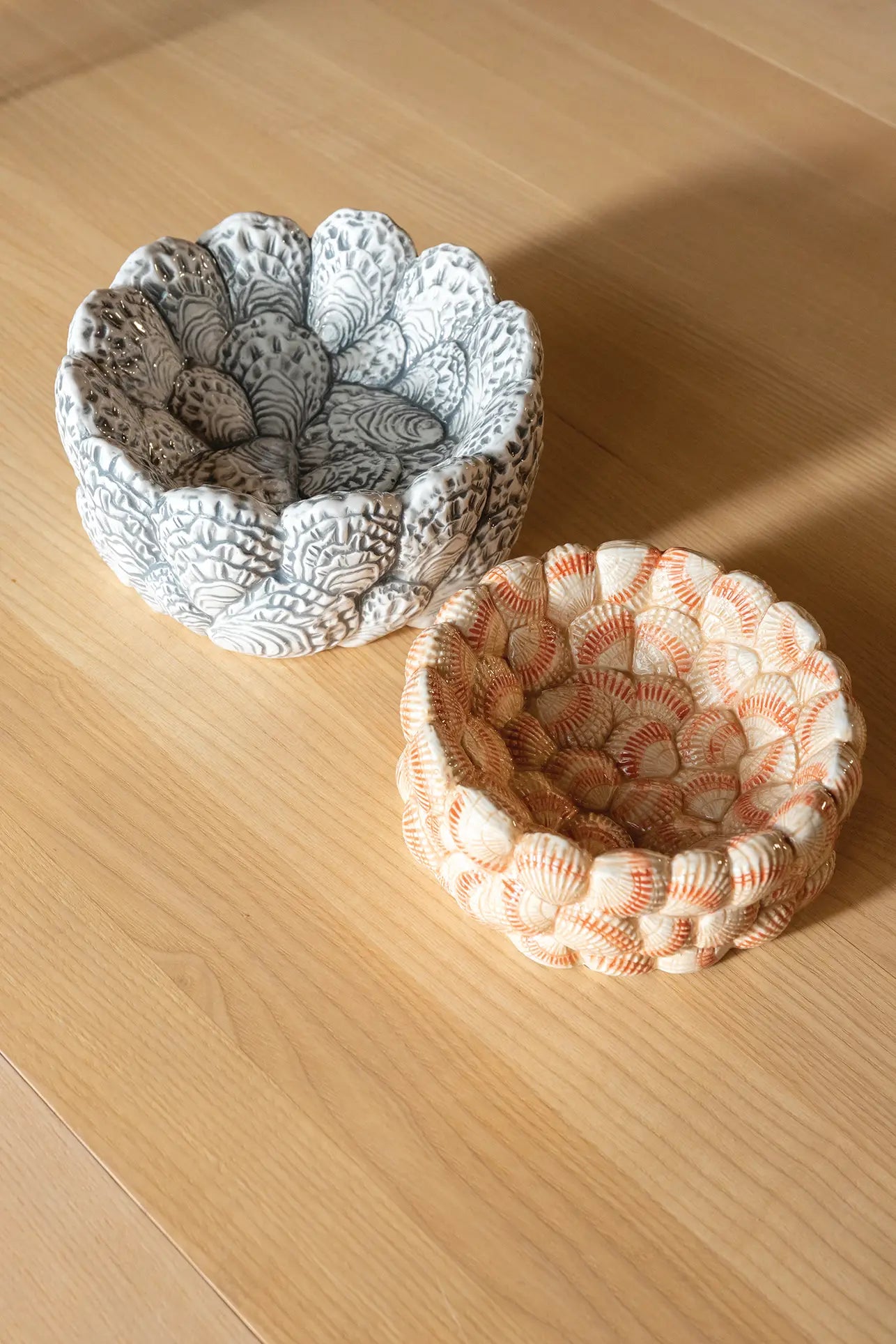 Two &klevering Oyster Bowls rest on a light wooden surface—one white with gray patterns, the other off-white with orange shell motifs—ideal for appetizers or oyster lovers.