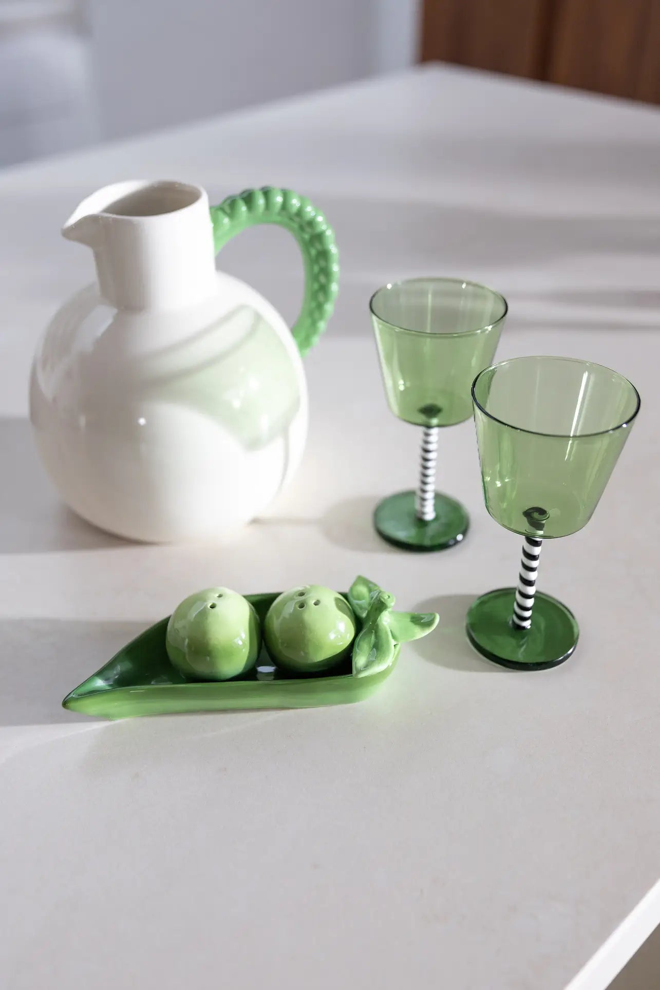 A white ceramic pitcher with a textured green handle, two green wine glasses with striped stems, and the &klevering Pea Salt & Pepper Set rest on a light kitchen counter.