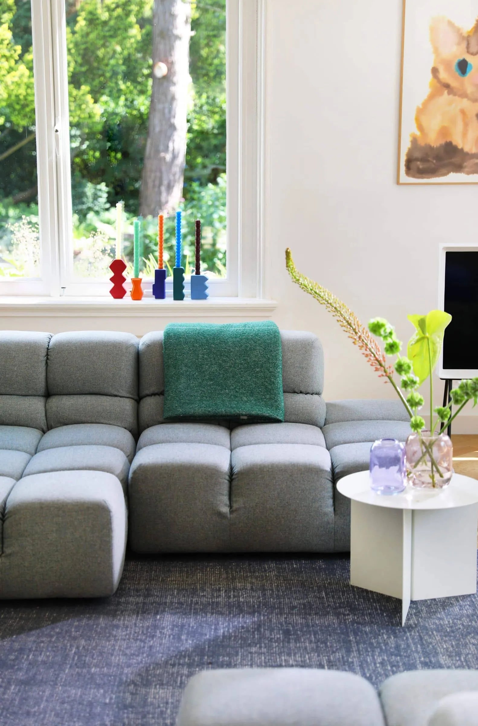 A modern living room with a grey tufted sectional, green throw blanket, white round table with vases and plants, &klevering Pebble Candle Set of 6 on the window sill, large windows, and a wall-mounted TV adds unique colour to the space.