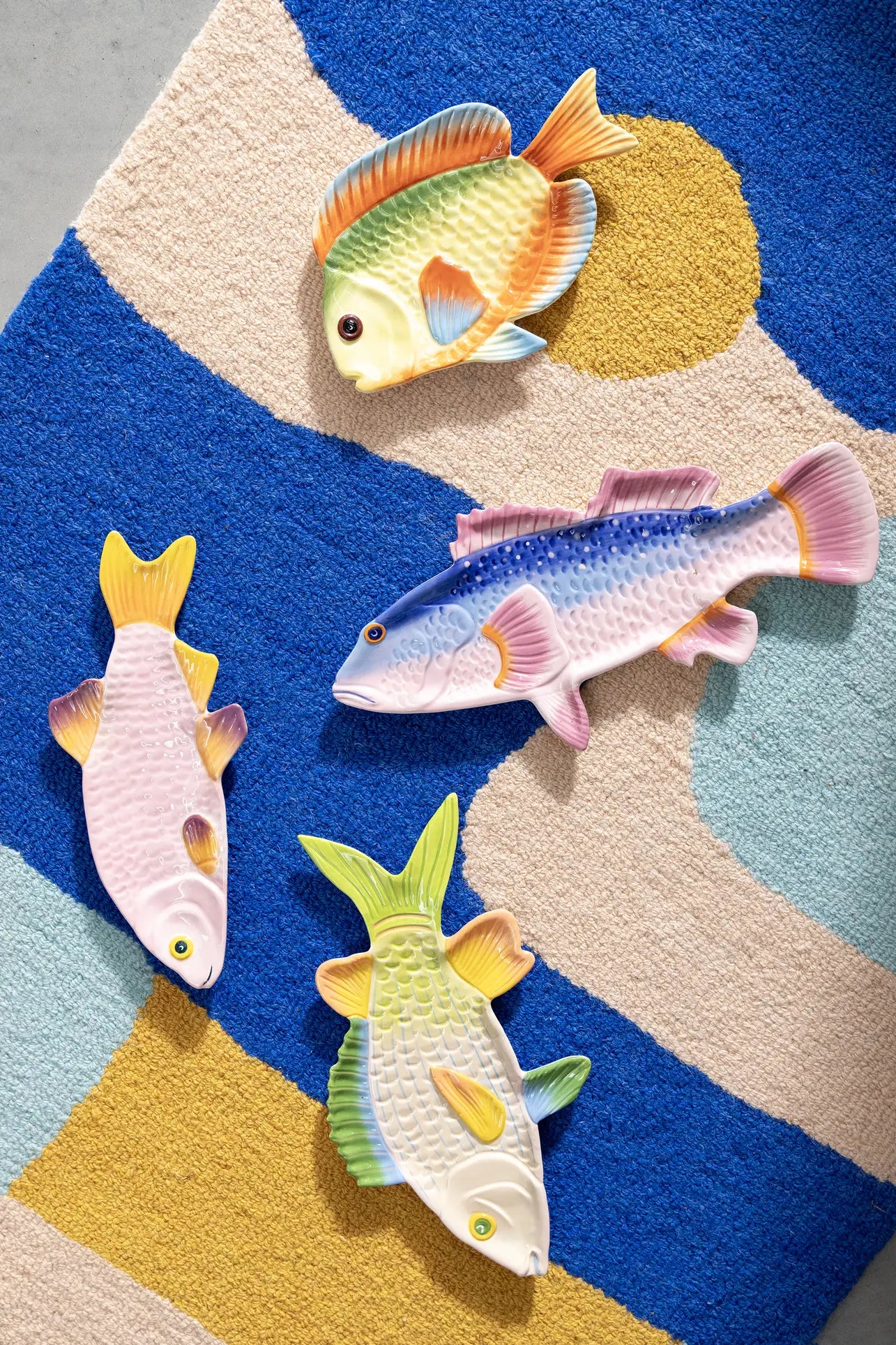 Four hand-painted ceramic dishes are displayed on a textured rug with blue, beige, and mustard yellow shapes. The set includes the &klevering Perch Fish Plate by &klevering, featuring vibrant details inspired by unique fish species.