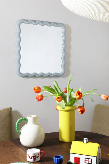 A modern dining table features a yellow vase of orange tulips, the &klevering Perle Jug, cups, and a small house ornament. A wavy-edged mirror above cushioned chairs adds elegance for perfect summer dining.