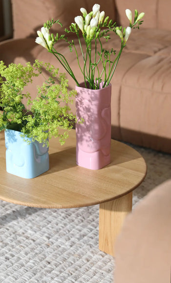 Two &klevering Puffy Vases—one light blue with green foliage, one pink with white flowers—sit on a round wooden coffee table, with a beige sofa and textured rug in the background.