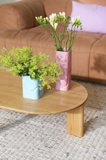 A wooden coffee table displays an &klevering Puffy Vase filled with vibrant flowers and a pink vase of white blooms. In the background, a beige couch with a lilac pillow rests on a textured rug.