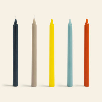 The &klevering Ripple Candle Set of 5 features tall black, beige, yellow, light blue, and orange candles with a modern vertical lined texture, all standing upright in a row against a light background.
