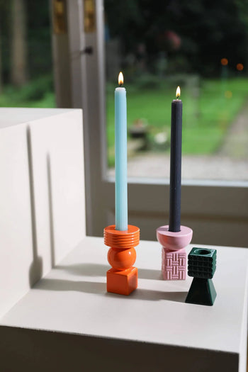The &klevering Ripple Candle, Set of 5 by &klevering features handcrafted candles with vertical lined textures, shown here in vibrant orange and pink beside a small green geometric holder by a window overlooking a blurred garden.