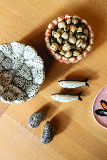 A light wooden table displays bowls of seashells, two ceramic fish, and hand-painted shell decor. The &klevering Sardine Salt & Pepper Set by &klevering and a pink dish with painted mussels enhance the marine-inspired arrangement.