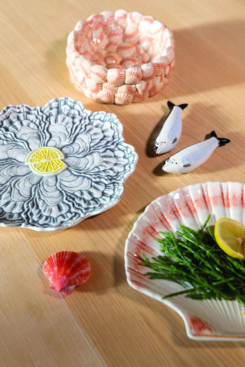 A decorative table setting with a blue floral plate, pink shell bowl, &klevering Sardine Salt & Pepper Set ceramic pieces, a scallop shell plate with greens and lemon, and a pink seashell on wood—evoking marine beauty.