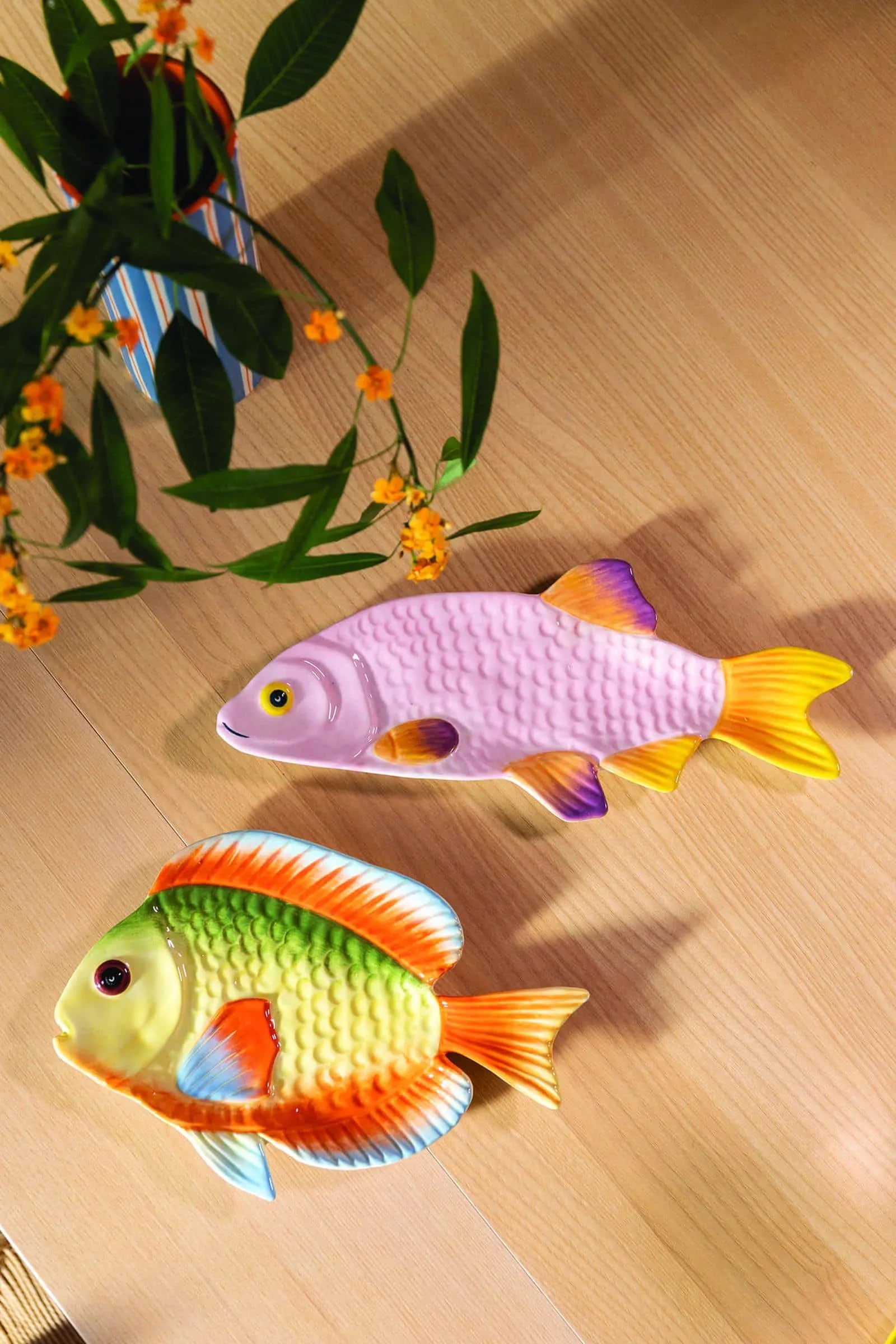 Two vibrant &klevering Snapper Fish Plates—one pink and yellow, the other green, orange, and yellow—brighten a light wooden table beside a vase with green leaves and orange flowers, making a playful centerpiece.