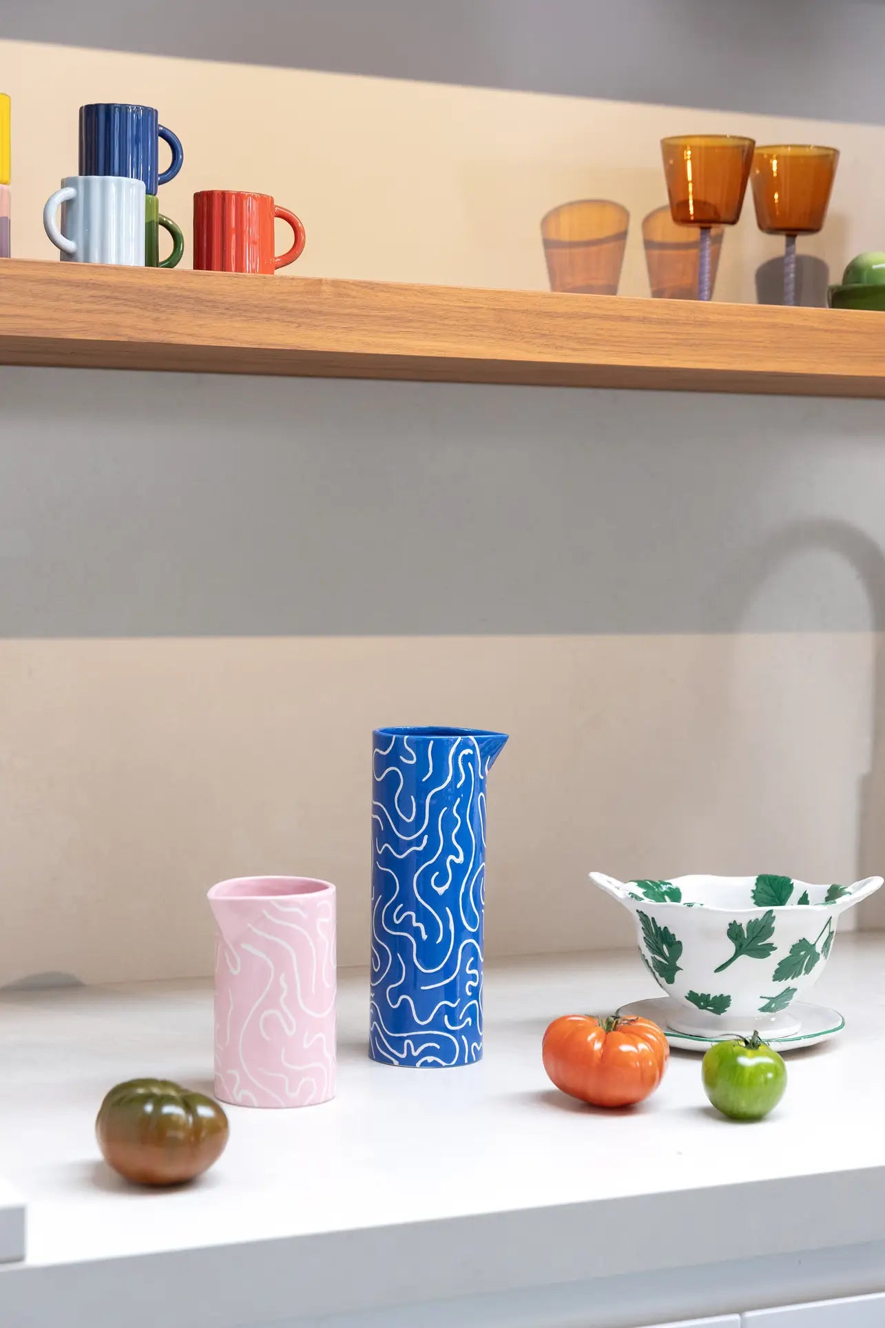 A white countertop features a pink and blue &klevering Soba Jug, a white bowl with green leaves, and three tomatoes. Above, colorful mugs with doodle prints and glasses sit neatly on a wooden shelf.