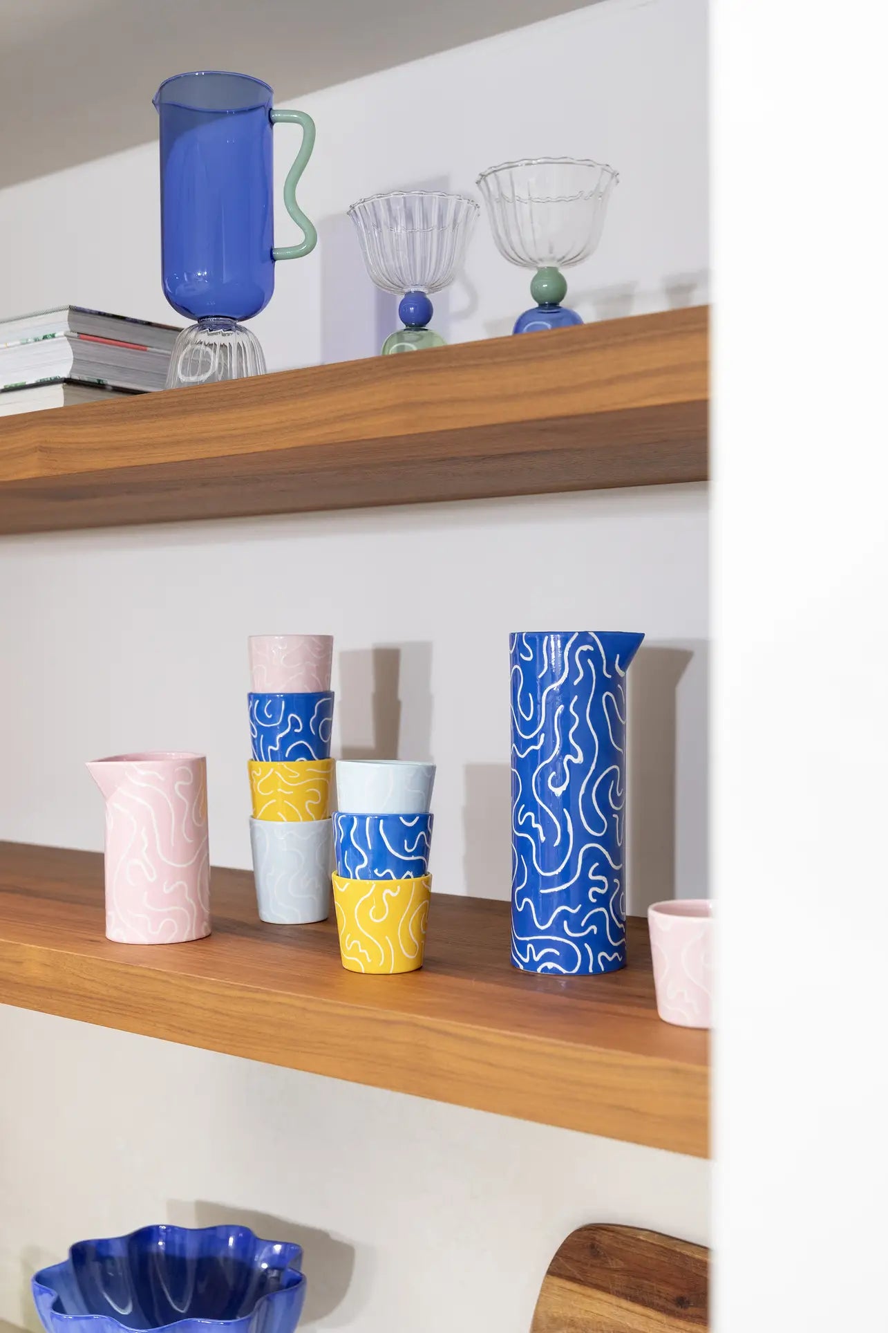 Cheerful doodle-print cups and pitchers in pink, yellow, blue, and white sit on a wooden shelf; above are glassware, stacked books, and an &klevering Soba Jug in bold blue.