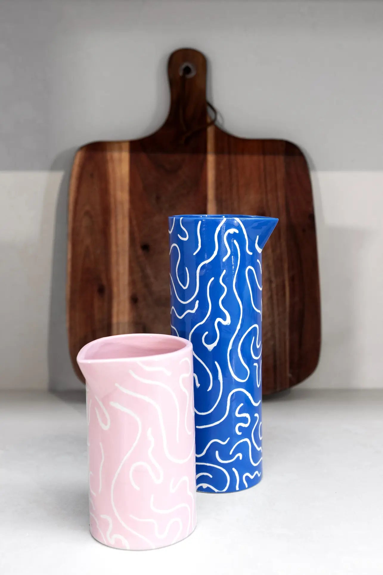 Two &klevering Soba Jugs in blue and pink with playful white squiggle designs sit before a wooden cutting board on a white countertop.