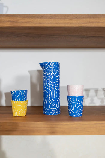 A vibrant ceramic drinkware set on a wooden shelf features the &klevering Soba Jug in bold blue with white swirls, matching blue and yellow cups, and two stacked pink cups from &klevering.