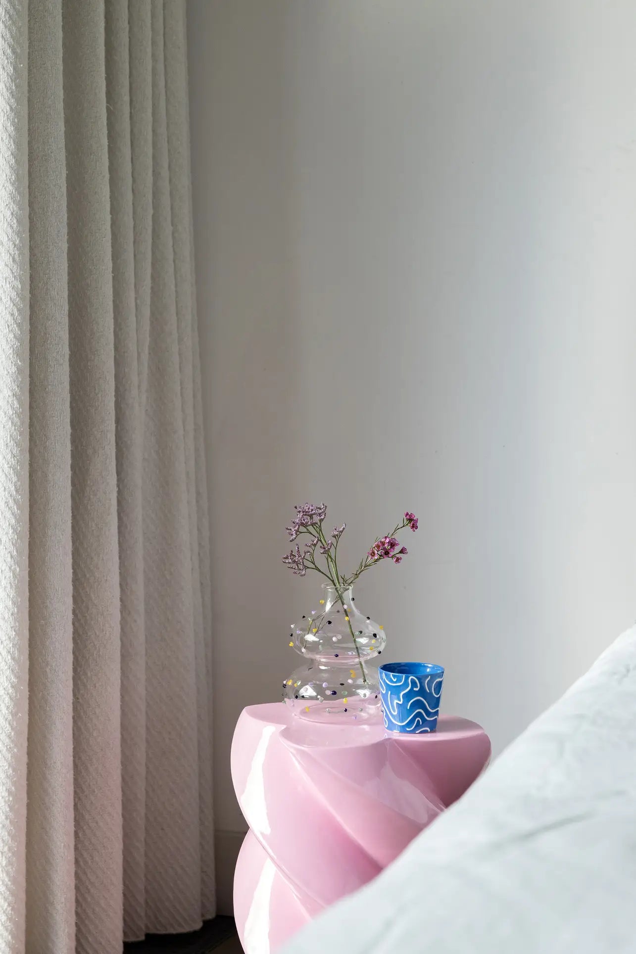 A modern, minimalist corner features a pink sculptural side table with a clear vase of delicate pink flowers and an &klevering Soba Mug Small from the set of 4. Vibrant colours pop, with a textured white curtain and bed edge in view.
