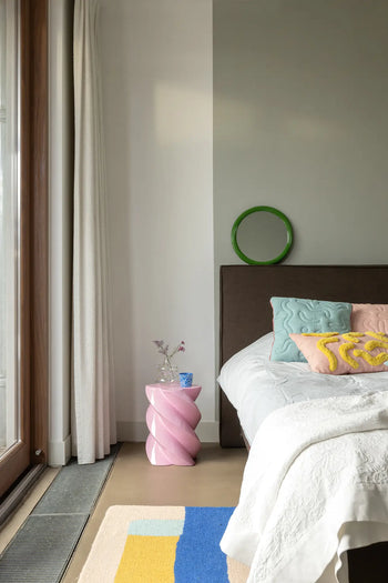 A modern bedroom with pastel quilted pillows, a white blanket, a pink spiral side table holding &klevering Soba Mug Small Set of 4 and flowers, green circular wall decor, floor-to-ceiling curtains, and a vibrant colorful rug.