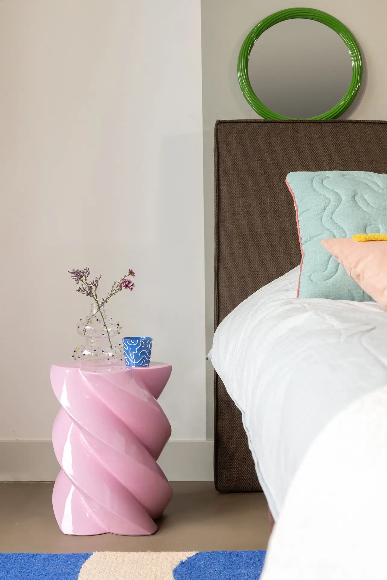 A modern bedroom with a brown upholstered bed, pastel bedding, and a pink spiral bedside table topped with a glass vase of flowers and &klevering Soba Mug Small set. Above hangs cheerful doodle art in a small round green mirror.