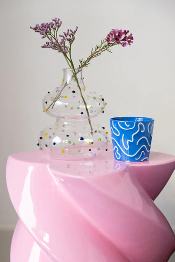 A cheerful clear glass vase with purple flowers sits next to a set of four &klevering Soba Mug Small mugs on a glossy pink, spiral-shaped table.