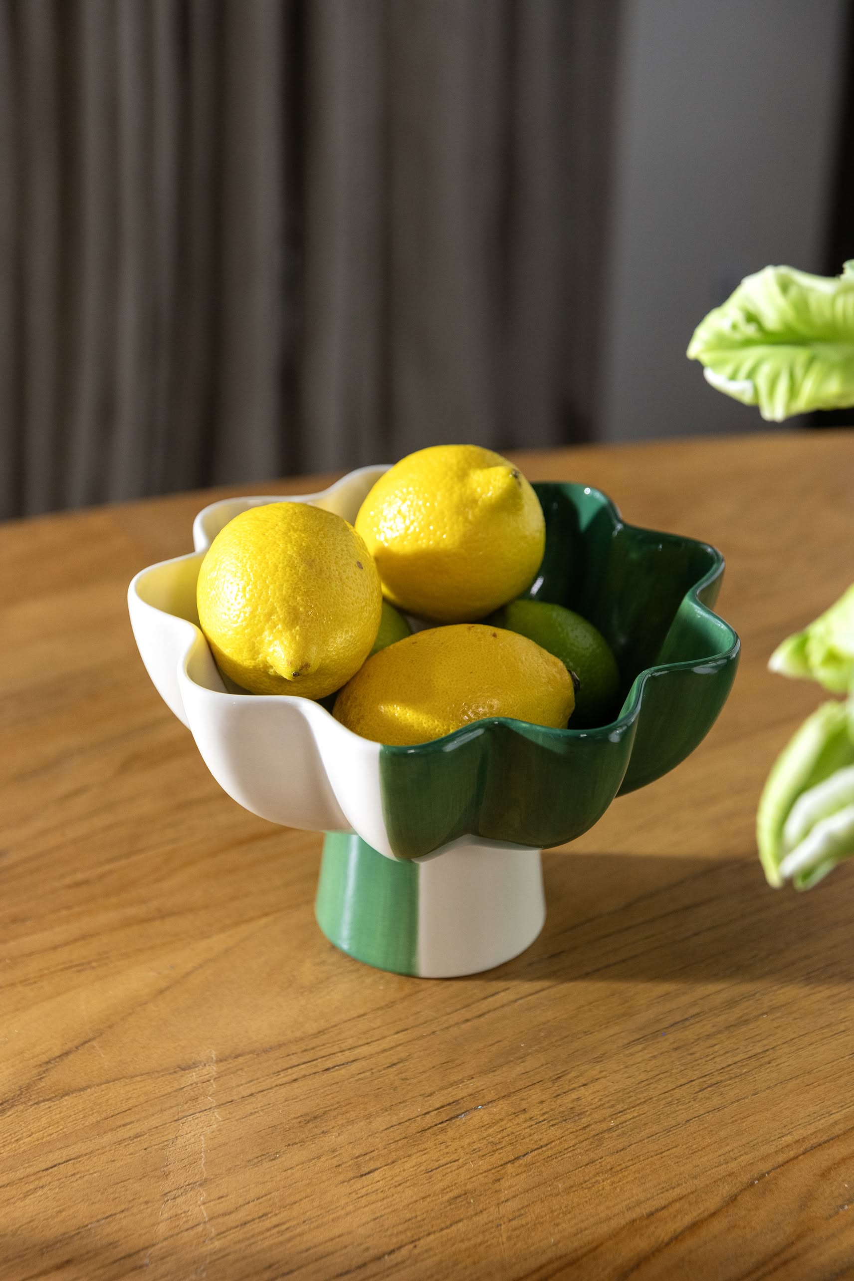 The &klevering Sun Bowl by &klevering, featuring green and white petals, displays four bright yellow lemons, making an eye-catching accent on a wooden table with a leafy plant to the right.