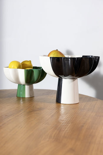 Two &klevering Sun Bowls—a green and white one with lemons and a black and white one with a pear—add a vibrant, modern accent to the minimalist scene on a wooden table against a plain background.