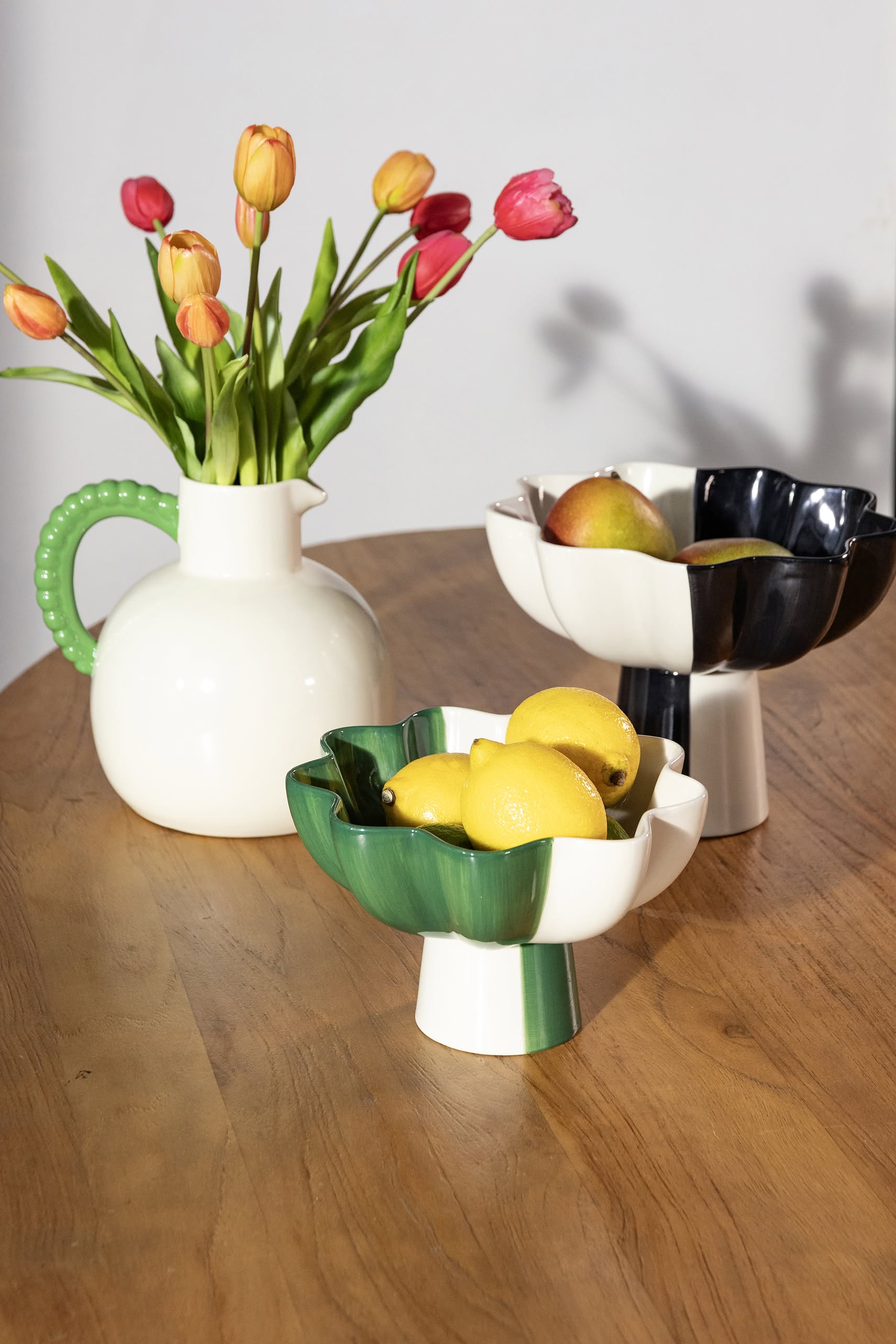 A white pitcher with pink tulips sits on a wooden table beside two &klevering Sun Bowls, one green and one black, filled with lemons and pears—an eye-catching accent from &klevering that brings vibrant modern style to any space.