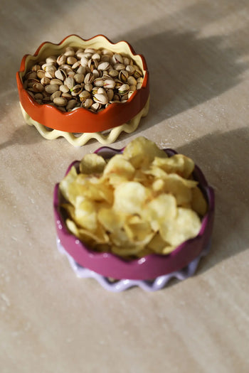 Two colorful &klevering Sway Trays with wavy rims sit on a beige surface: the purple tray holds potato chips and the orange tray offers pistachios—perfect for stylish storage or as a unique centerpiece.