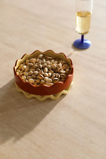The &klevering Sway Tray ceramic bowl by &klevering, filled with pistachios, rests on a beige surface beside a blue-stemmed glass, adding modern style and practical storage to any setting.