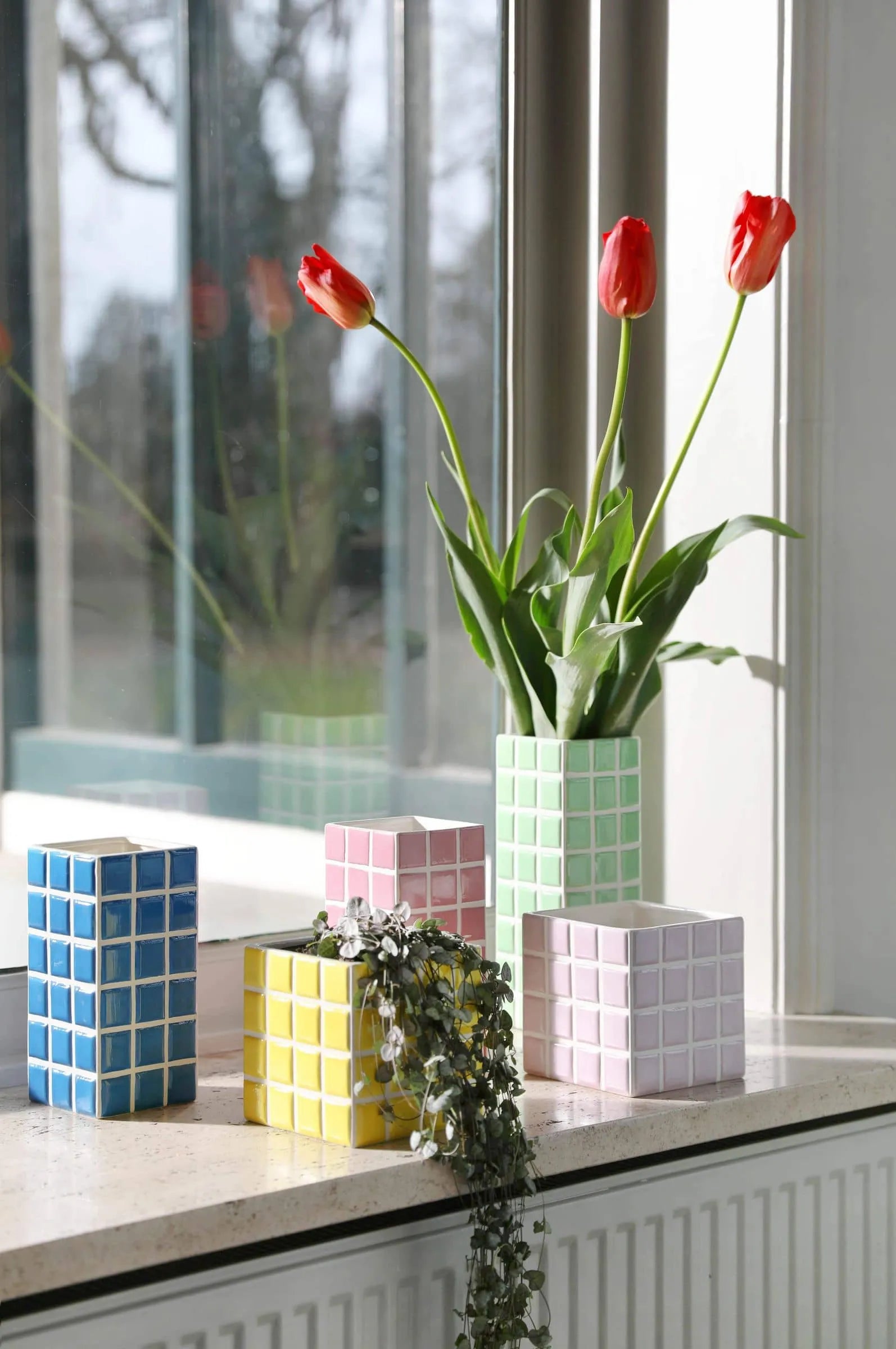 A green checkered planter with red tulips sits on a windowsill, surrounded by four pastel &klevering Tiled Planter Lilac vases in blue, yellow, pink, and white hues, plus a small trailing plant. The window reflects this vibrant display.