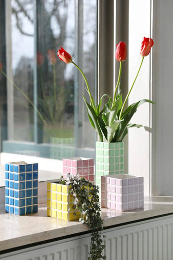A green checkered planter with red tulips sits on a windowsill, surrounded by four pastel &klevering Tiled Planter Lilac vases in blue, yellow, pink, and white hues, plus a small trailing plant. The window reflects this vibrant display.