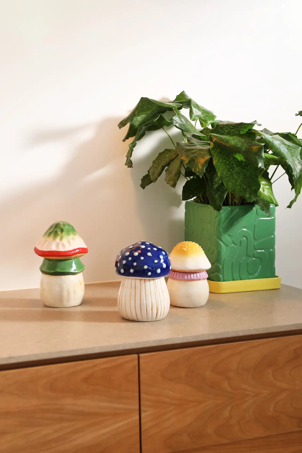 Three colorful ceramic mushroom ornaments sit on a wooden cabinet beside the &klevering Tribe Planter (Green) by &klevering, featuring a large-leafed plant in a geometric pot with drainage hole, all set against a plain white wall.