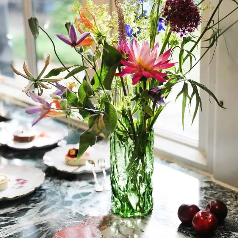 An &klevering Trunk Vase by &klevering, filled with vibrant wildflowers, stands on a marble table near a window, surrounded by plates of desserts and several plums.