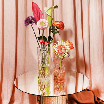 Two &klevering Trunk Vases by &klevering, filled with assorted pink, purple, orange, and green flowers, are arranged on a round mirrored table in front of pinkish-beige curtains.