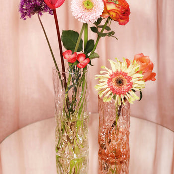 The &klevering Trunk Vase by &klevering, in clear and pink glass, displays vibrant pink, orange, and yellow flowers on a reflective surface, softly framed by a pale pink curtain.