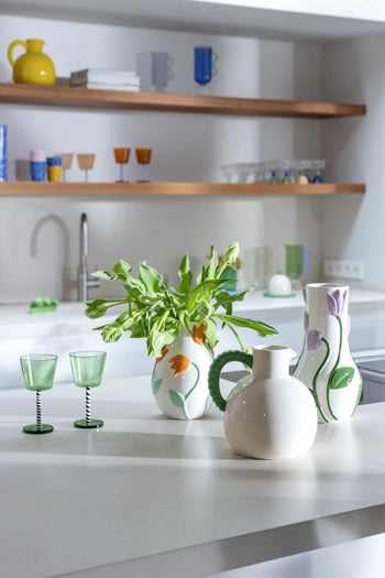 A modern, Netherlands-inspired kitchen features a white countertop with three decorative vases, including the &klevering Tulip Vase, Small with green tulips. Two green glasses are nearby, and wooden shelves above hold cups and glasses.
