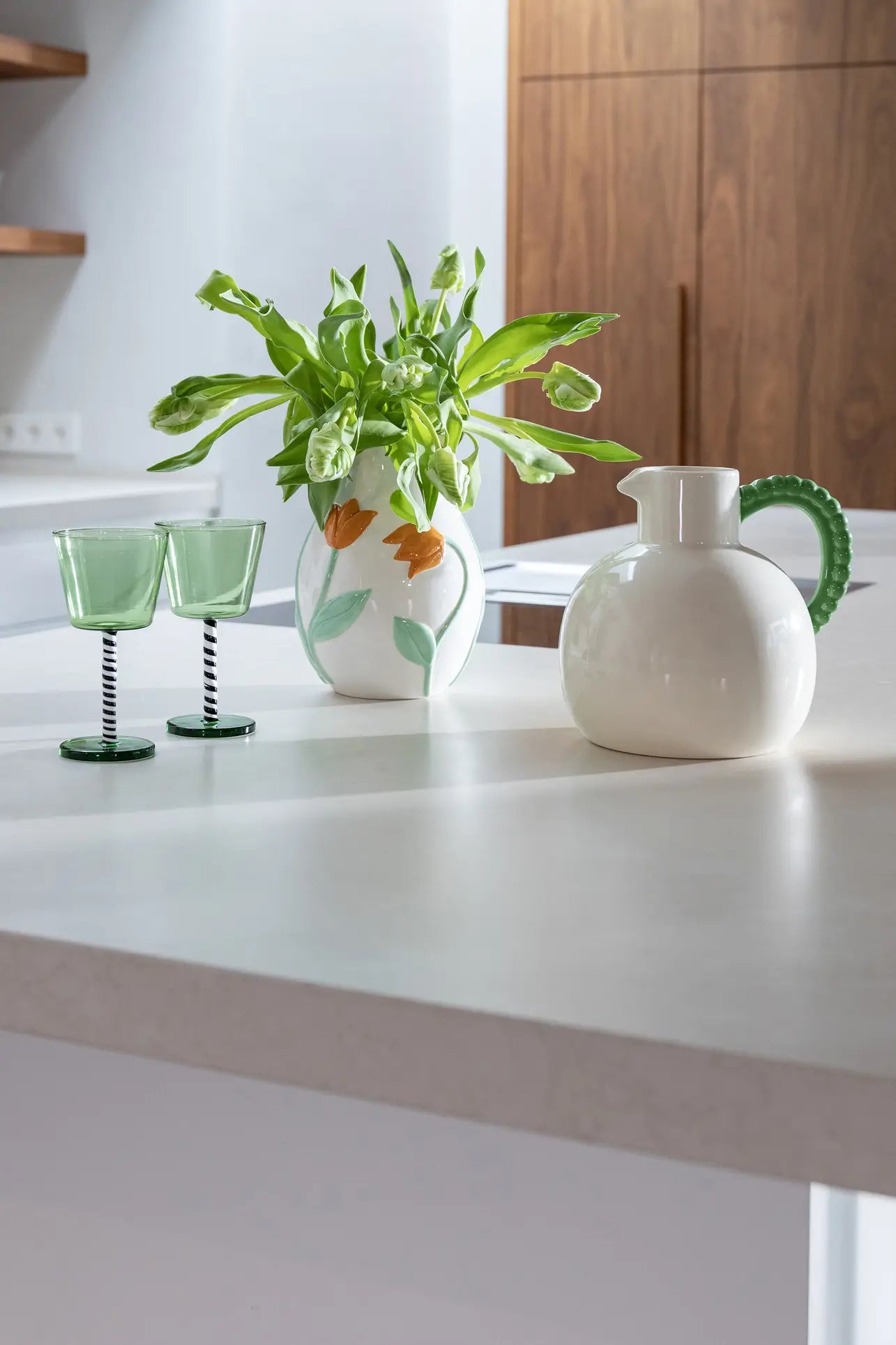 A white kitchen countertop displays two green glasses, the &klevering Tulip Vase, Small with leafy plants, and a round white pitcher with a green handle. Wood cabinets in the background create a Netherlands-inspired ambiance.