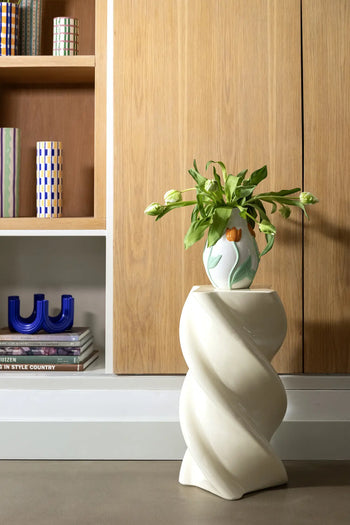A green plant in the &klevering Tulip Vase, Small sits on a white twisted pedestal, while books and decor nearby are arranged on shelves against light wood-paneled cabinets for a Netherlands-inspired touch.