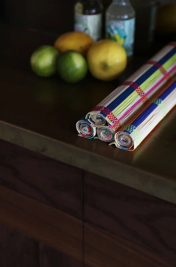A set of three colorful &klevering Umami Placemats rests on a dark wooden countertop, with blurred lemons, limes, and bottles in the background.