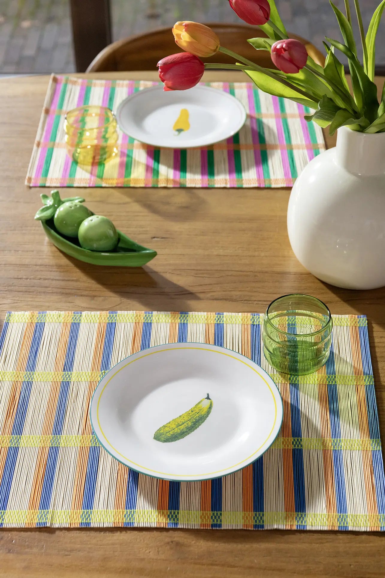 A wooden dining table is set with &klevering Umami Placemat Set of 4, plates with painted vegetables, green glasses, a white vase of red and yellow tulips, and a ceramic pea pod dish. Sunlight streams in, brightening this charming setup.
