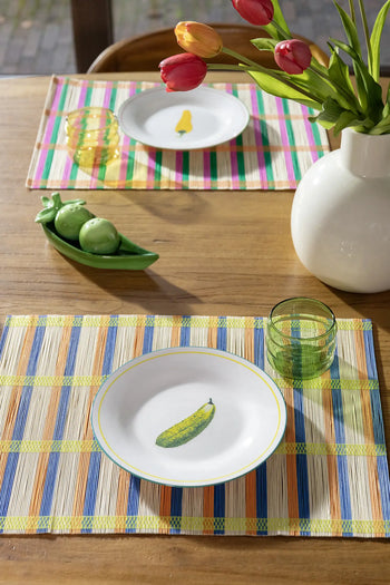 A wooden dining table is set with &klevering Umami Placemat Set of 4, plates with painted vegetables, green glasses, a white vase of red and yellow tulips, and a ceramic pea pod dish. Sunlight streams in, brightening this charming setup.