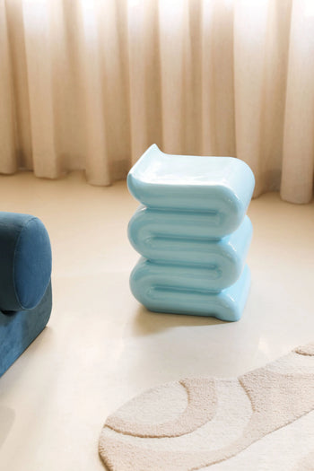 The &klevering Whip Pillar - Blue, a pastel wavy sculptural side table by &klevering, stands on a beige floor beside a plush blue chair and textured cream rug, forming a cozy reading nook with beige curtains behind.