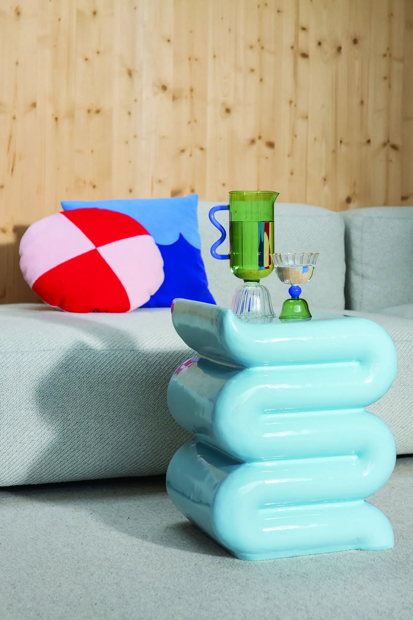 A modern living room with a light gray sofa and the &klevering Whip Pillar - Blue from &klevering as a colorful accent. A pastel side table holds a green pitcher, glass, and bowl, while the wooden wall backdrop adds to this cozy reading nook.