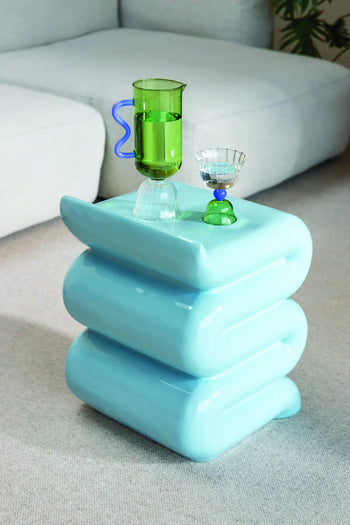 The &klevering Whip Pillar - Blue by &klevering, a small glossy pastel side table shaped like stacked cushions, sits on carpet serving as a portable drinks counter, topped with a green glass pitcher and cup, with a light sofa behind.