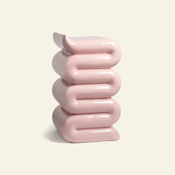 The &klevering Whip Pillar - Pink by &klevering, with its shiny pastel surface and stacked, folded design reminiscent of curved ribbons or soft cushions, stands against a plain off-white background.